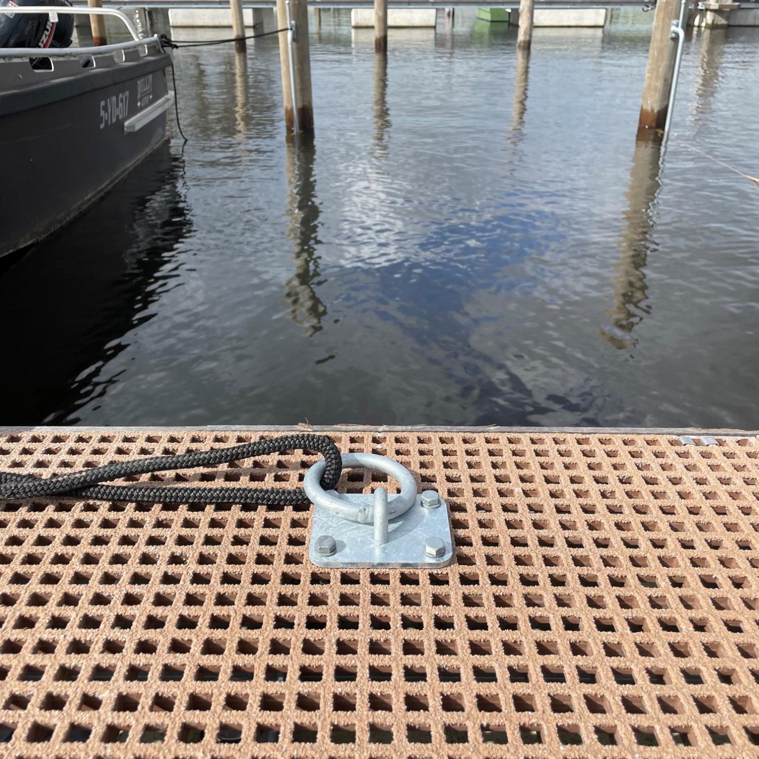  Een thermisch verzinkte afmeerring bevestigd op een open roostervloer van een steiger, met een aangemeerd schip en een rustige haven op de achtergrond.