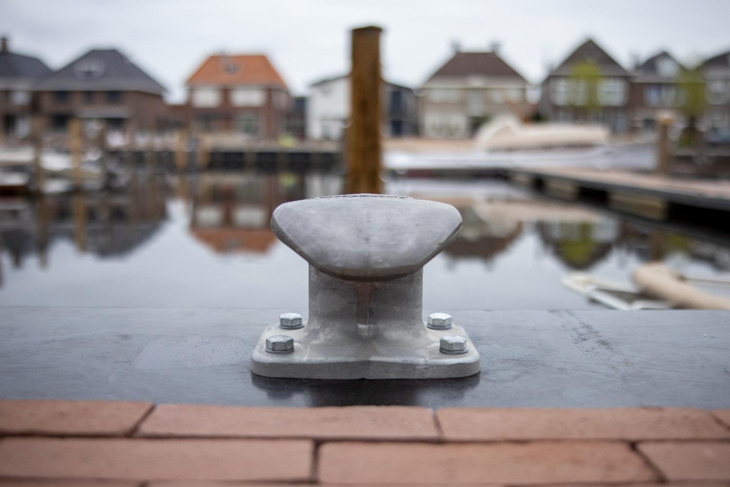 Een Walcon MH5 bolder bevestigd op een kade met zicht op een haven en woningen op de achtergrond.