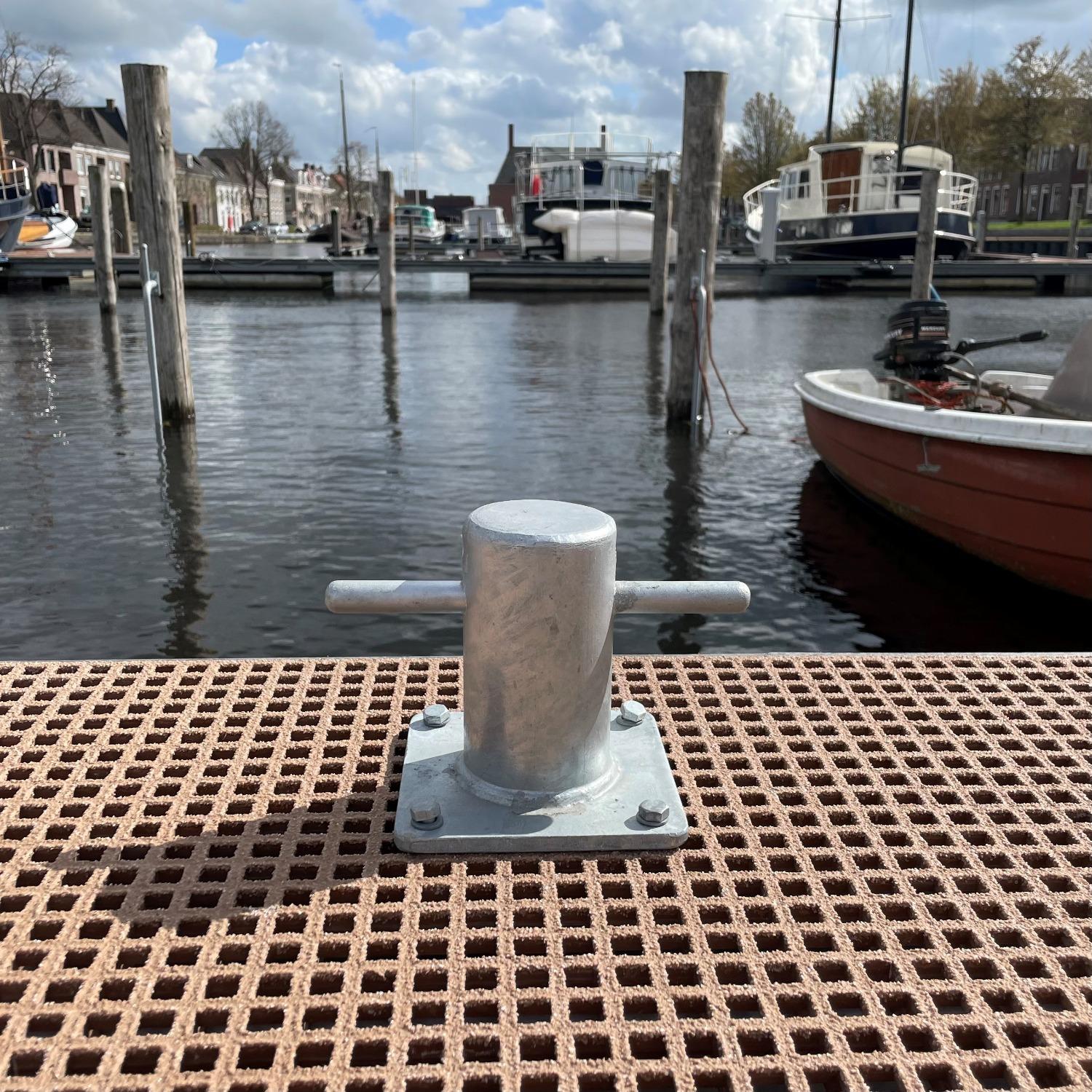  en stevige thermisch verzinkte bolder gemonteerd op een open roostervloer van een steiger, met een haven en aangemeerde boten op de achtergrond.