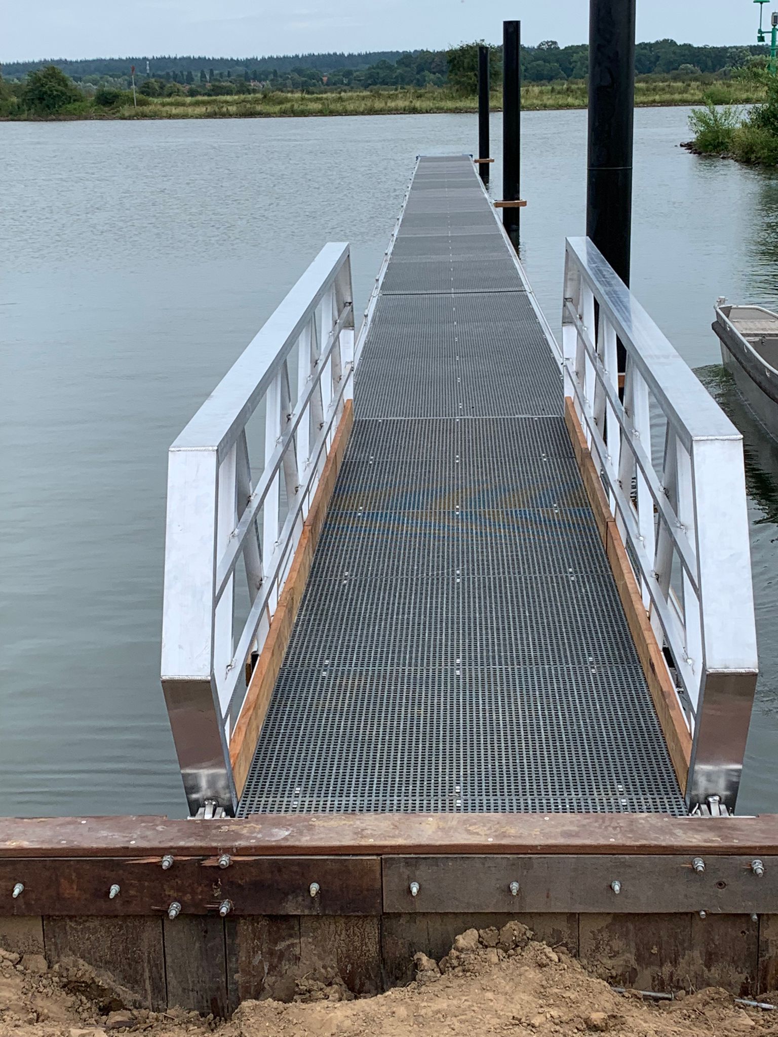 Ein detailliertes Bild eines Aluminiumweges mit Holzkanten und GVK -Grills, die an den Ufern eines Wassers aufgestellt sind. Die Brücke führt zum Wasser und wird von schwarzen Multi -Poles unterstützt. Im Hintergrund sind ein grünes Ufer und ein Flussleuger unter einer bewölkten Luft sichtbar.