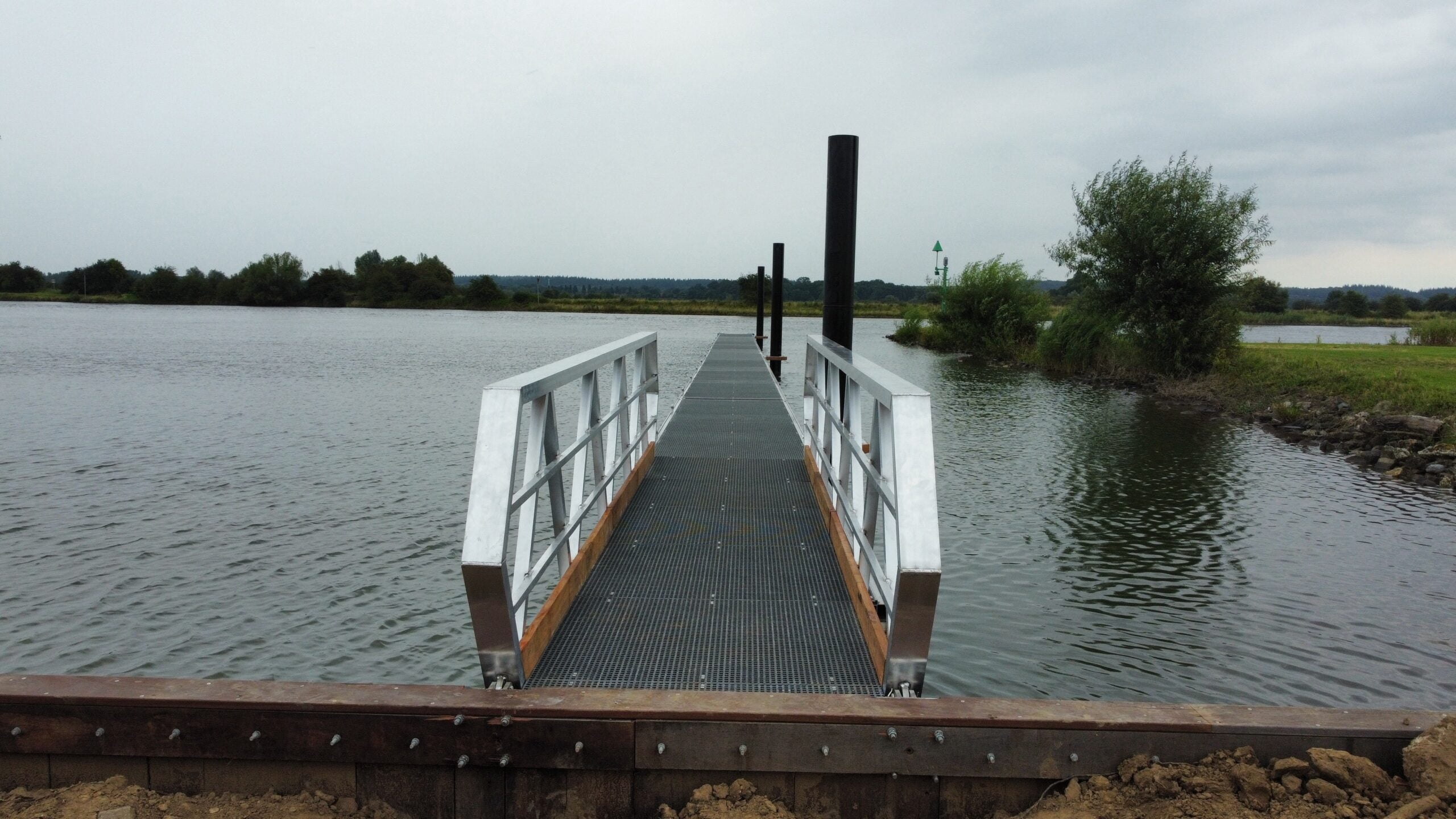 Ein Aluminiumweg mit Holzkanten und GVK -Grills, die an den Ufern eines ruhigen Wassers gelegt werden. Die Brücke führt zum Wasser und wird von schwarzen Multi -Poles flankiert. Im Hintergrund sind grüne Landschaft, Sträucher und ein Flussleuger unter einer bewölkten Luft sichtbar.
