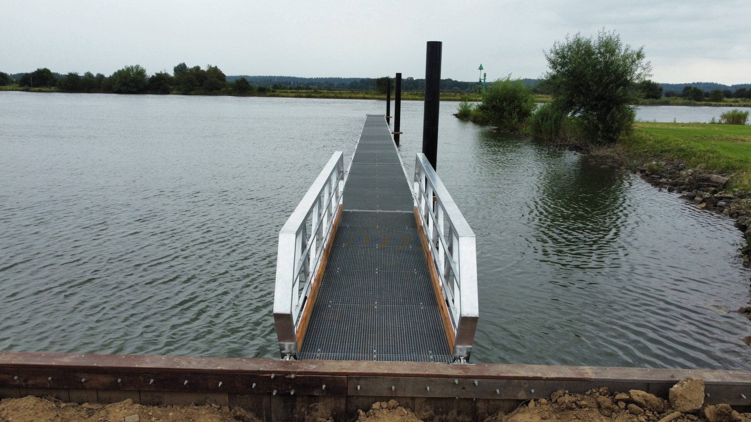 Obere Blick auf einen Aluminiumweg mit Holzkanten und GVK -Grills, der sich am Ufer eines Wassers befindet. Die Brücke wird von schwarzen Multi -Polen unterstützt und führt zum Wasser, wobei eine grüne Umgebung und ein im Hintergrund im Hintergrund sichtbarer Flussleuchtfeuer unter einer bewölkten Luft sichtbar sind.