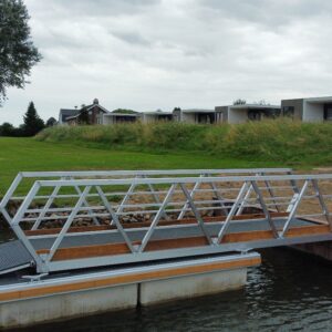 Seitenansicht eines Aluminiumweges mit Holzfinish, der über einem Wasserspiel platziert ist. Die Brücke verfügt über ein modernes geometrisches Design und ruhen Sie sich auf einer konkreten Fundament aus. Im Hintergrund befindet sich ein grünes Grasflugzeug mit Häusern und einem historischen Kamin, der unter einem bewölkten Himmel sichtbar ist.