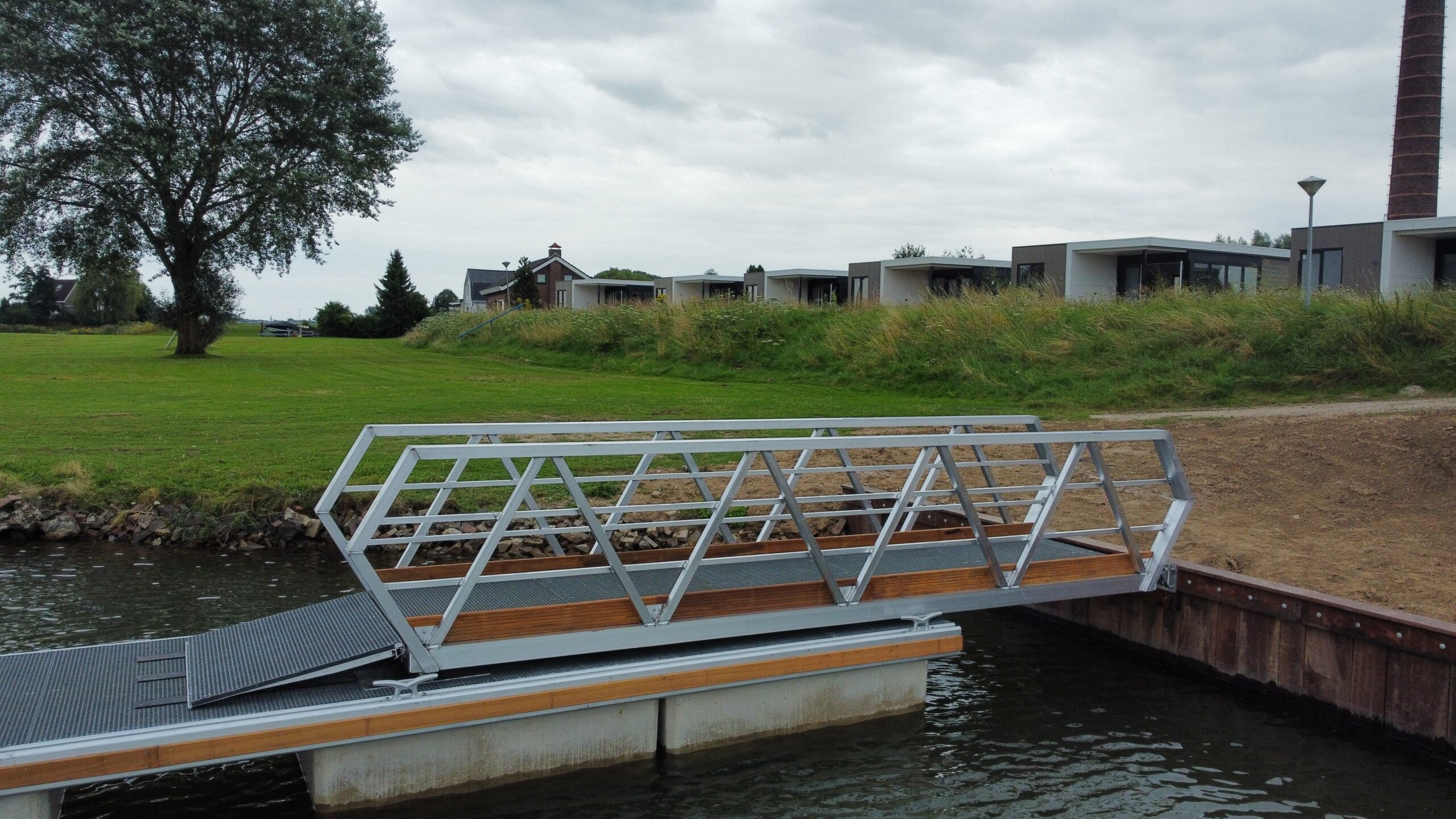 Seitenansicht eines Aluminiumweges mit Holzfinish, der über einem Wasserspiel platziert ist. Die Brücke verfügt über ein modernes geometrisches Design und ruhen Sie sich auf einer konkreten Fundament aus. Im Hintergrund befindet sich ein grünes Grasflugzeug mit Häusern und einem historischen Kamin, der unter einem bewölkten Himmel sichtbar ist.