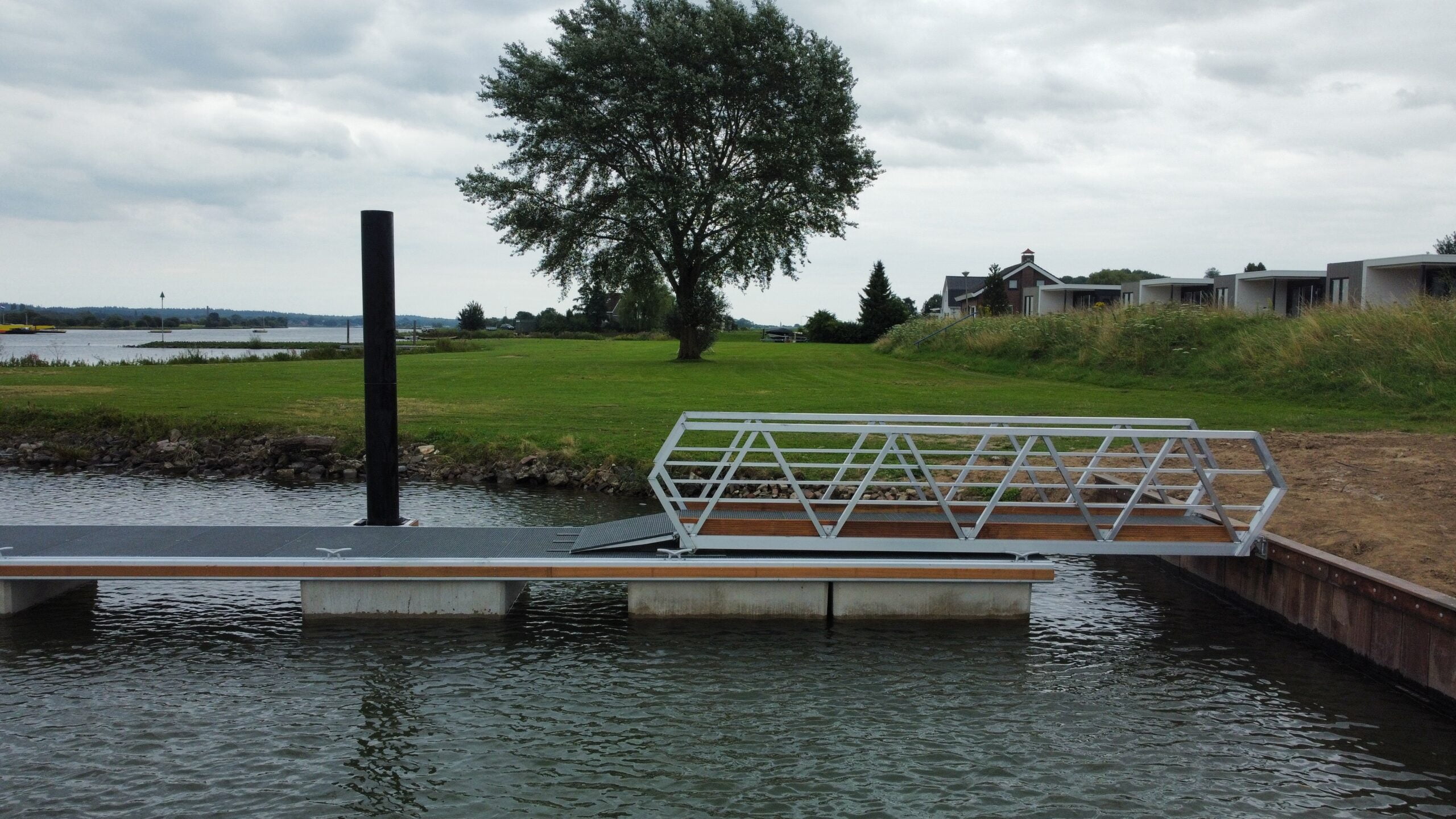 Moderner Steg mit einem Metallweg und einer Holzplattform an einer Uferpromenade, umgeben von Gras und Bäumen.