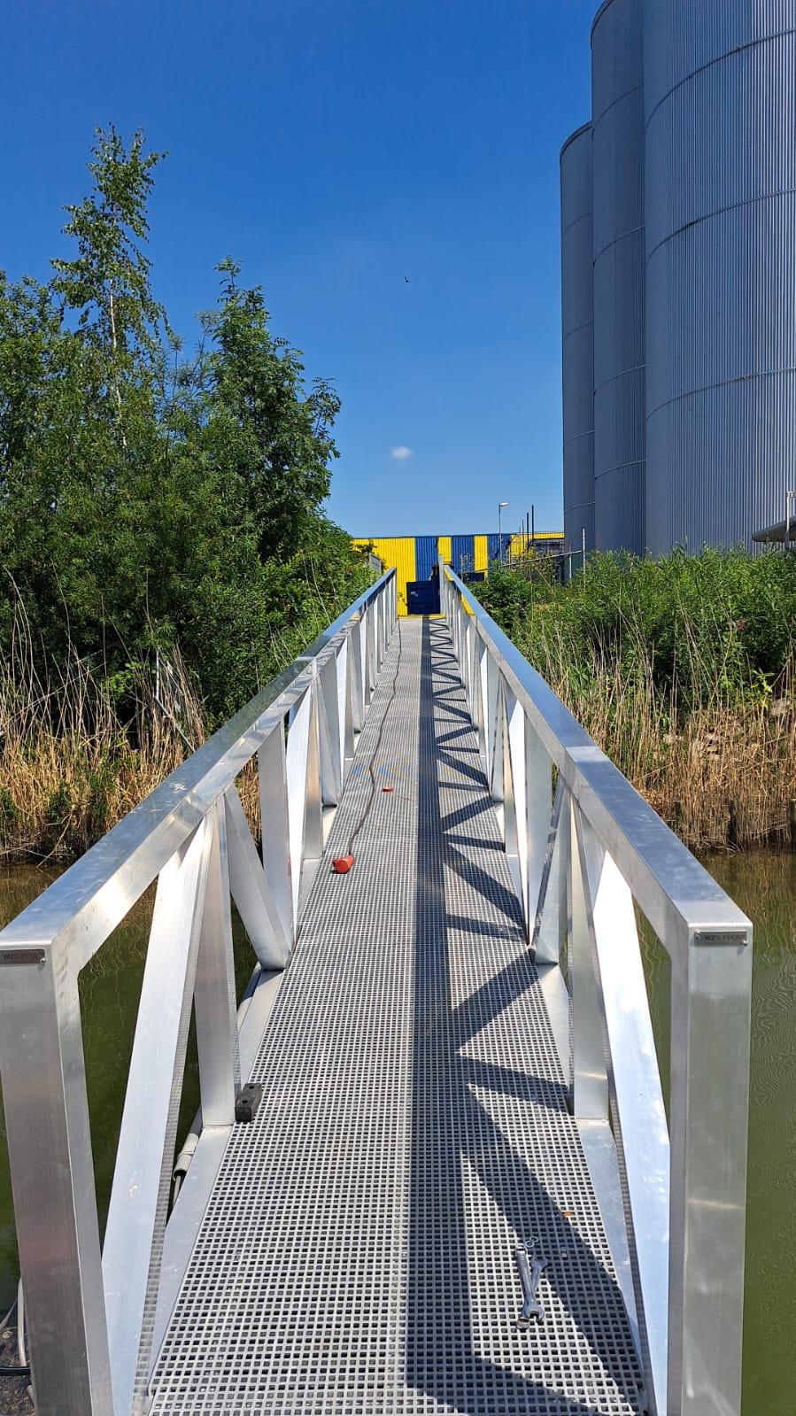 Ein Aluminiumweg über einem Wasser mit grüner Vegetation und Schilf. Die Brücke hat ein robustes Zeitplandesign und bietet einen sicheren Durchgang. Im Hintergrund sind industrielle Silos und eine gelbblaue Struktur gegen einen klaren blauen Himmel sichtbar.