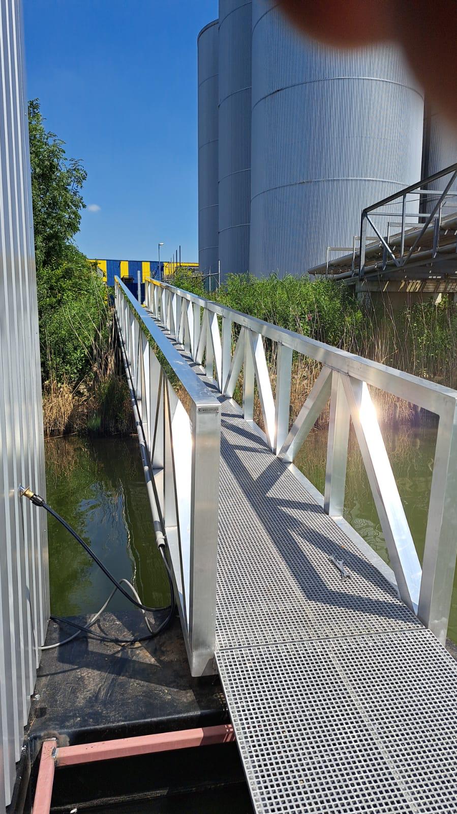 Seitenansicht eines Aluminiumweges über einem Wasserspiel mit einem robusten Netzdesign und stabilen Handläufen. Die Brücke ist von grüner Vegetation und Schilf umgeben, mit industriellen Silos und einer gelbblauen Struktur, die im Hintergrund unter einem klaren blauen Himmel sichtbar ist.