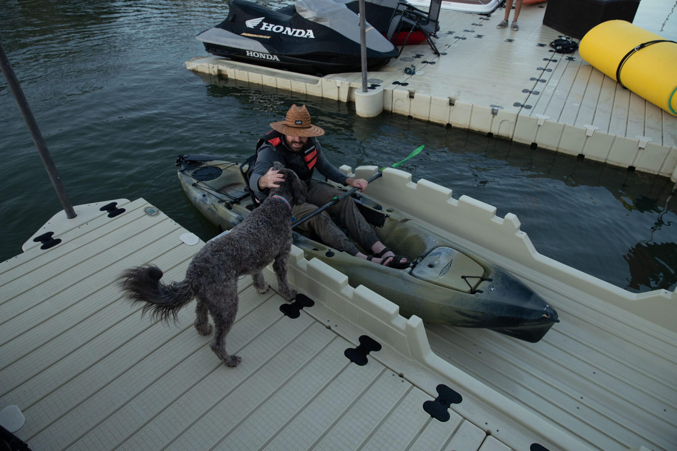 Een man in een kajak op een EZ Dock kajakplatform, terwijl hij zijn hond op de steiger aait. Op de achtergrond liggen jetski's in het water.